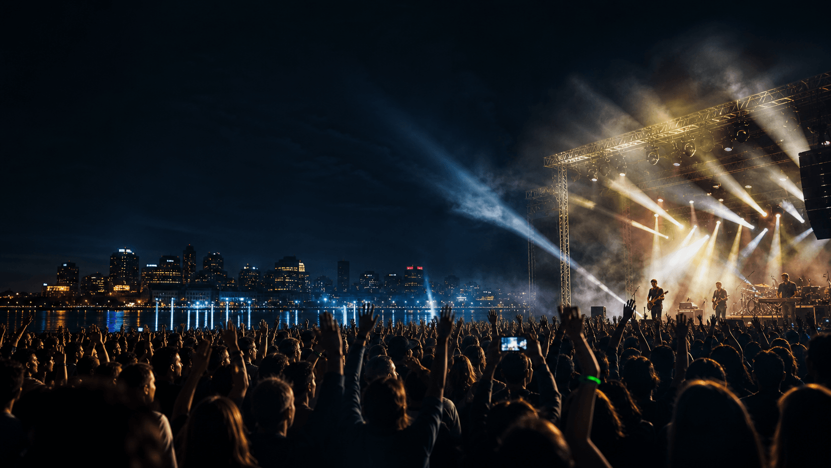 Outdoor night concert with crowd, riverfront city lights, and sweeping stage beams